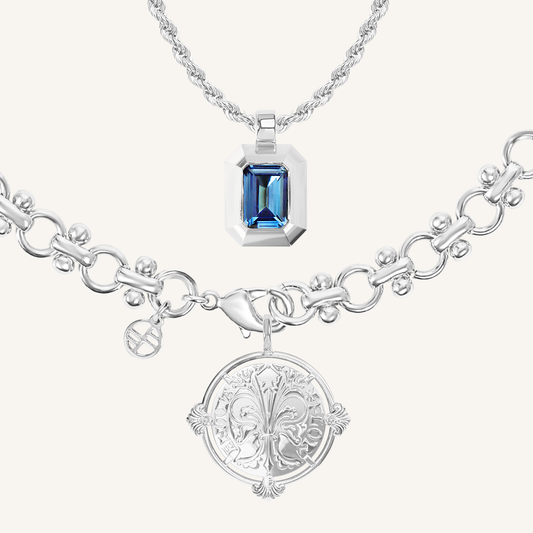 silver chain necklace inspired by Eiffel tower architecture with silver pendant featuring intricate patterns inspired by Palace of Versailles next to rectangular charm featuring deep blue cubic zirconia on white background