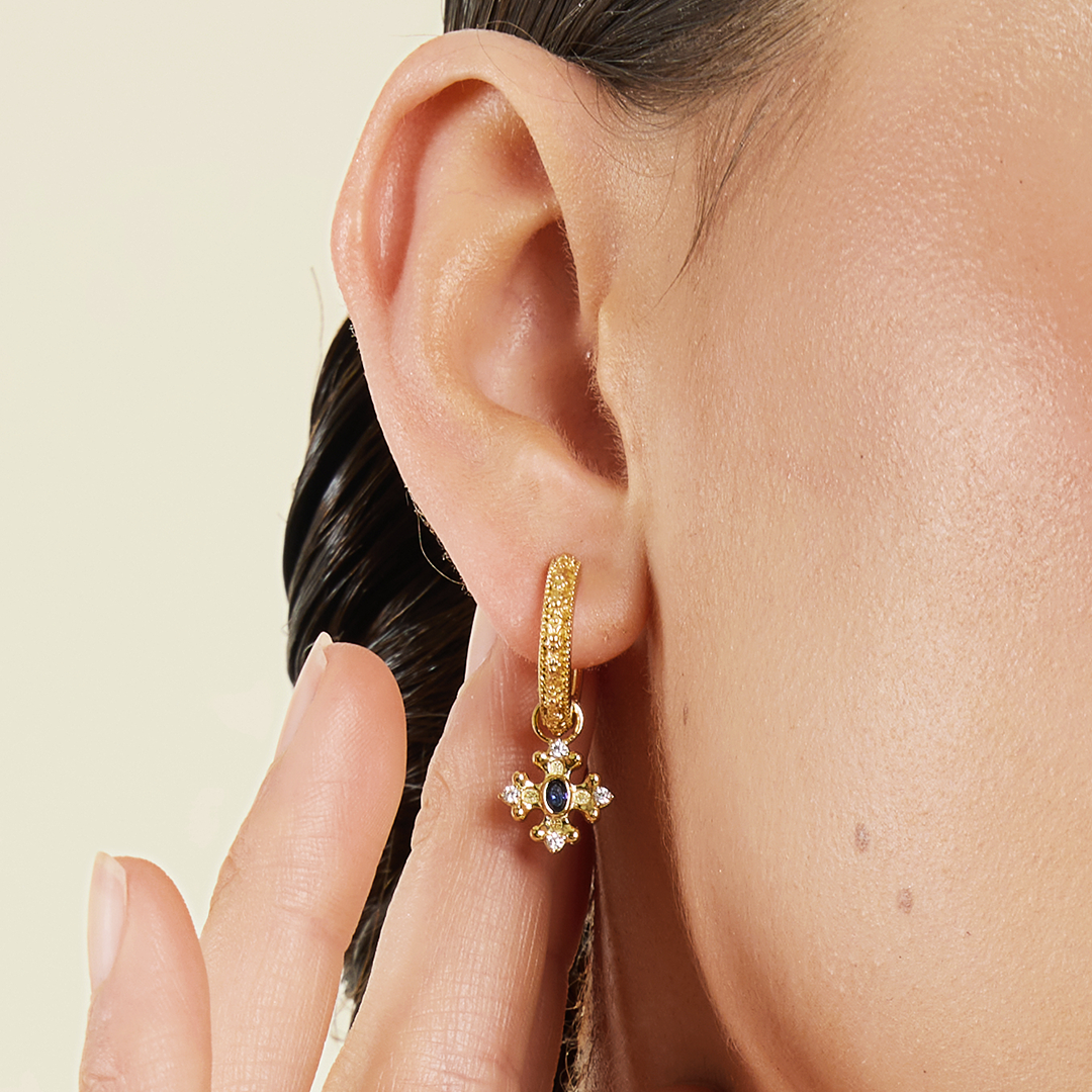 gold charm that features the intricate fleur-de-lis details found in the iconic Palace of Versailles accompanied by the deep blue stone on gold hoops on the ear close up