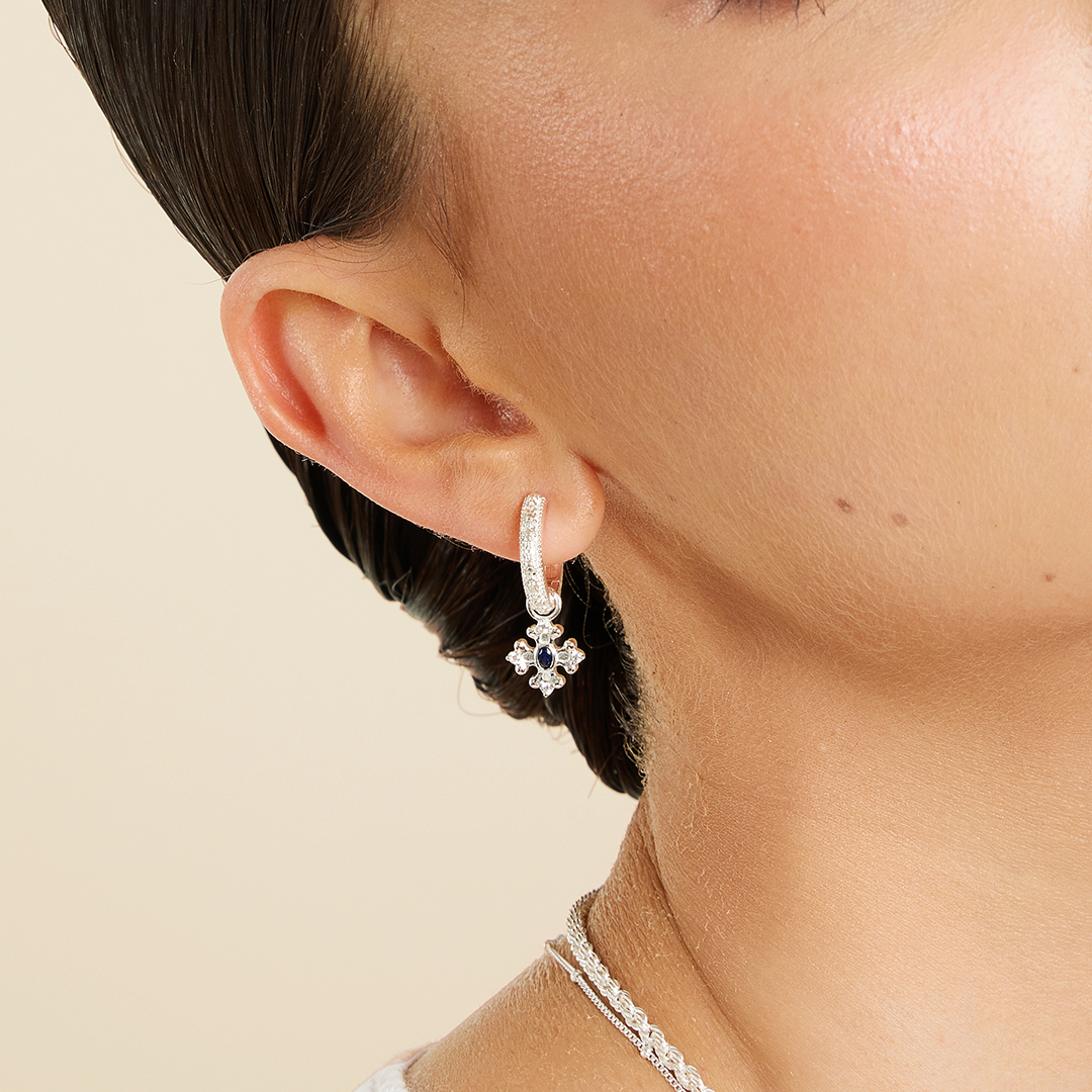 silver charm that features the intricate fleur-de-lis details found in the iconic Palace of Versailles accompanied by the deep blue stone on gold hoops close up on ear 