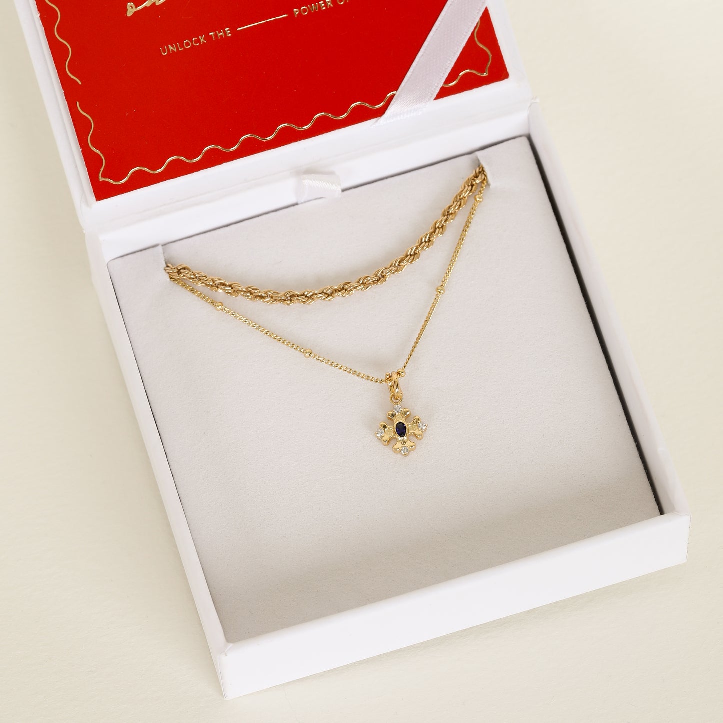 gold charm that features the intricate fleur-de-lis details found in the iconic Palace of Versailles accompanied by the deep blue stone on bobble chain next to rope chain in white box 