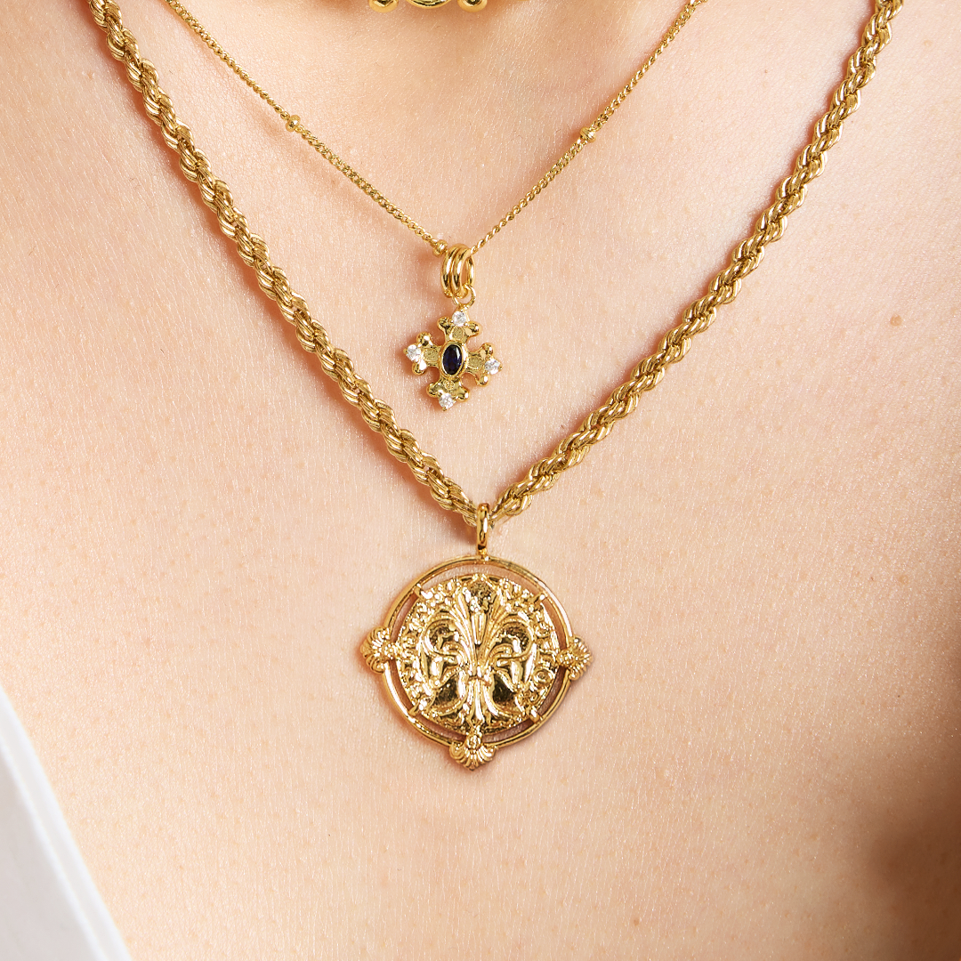 gold pendant featuring intricate patterns inspired by Palace of Versailles on gold rope chain paired with gold bobble chain with gold and blue charm on neck