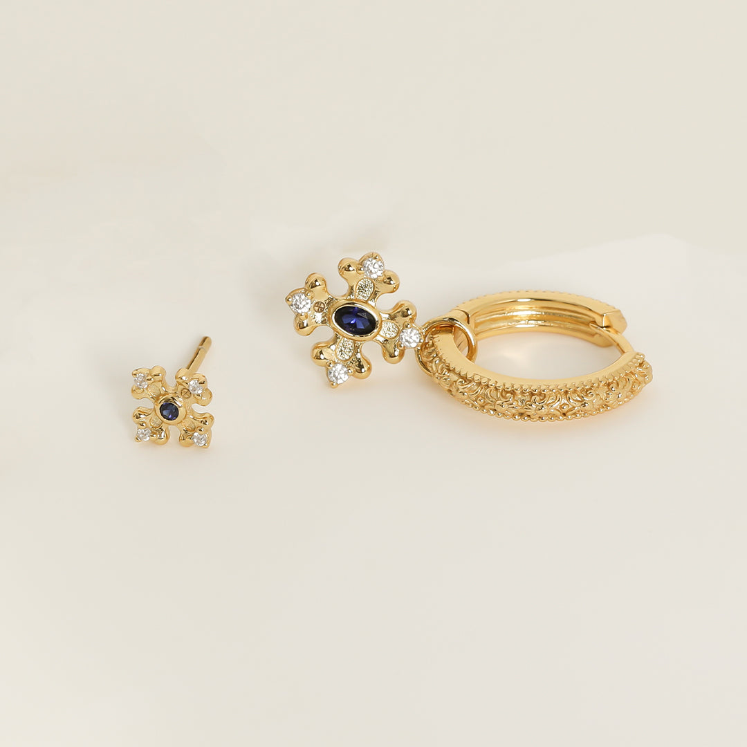 gold charm that features the intricate fleur-de-lis details found in the iconic Palace of Versailles accompanied by the deep blue stone on gold hoops  next to stud on white background