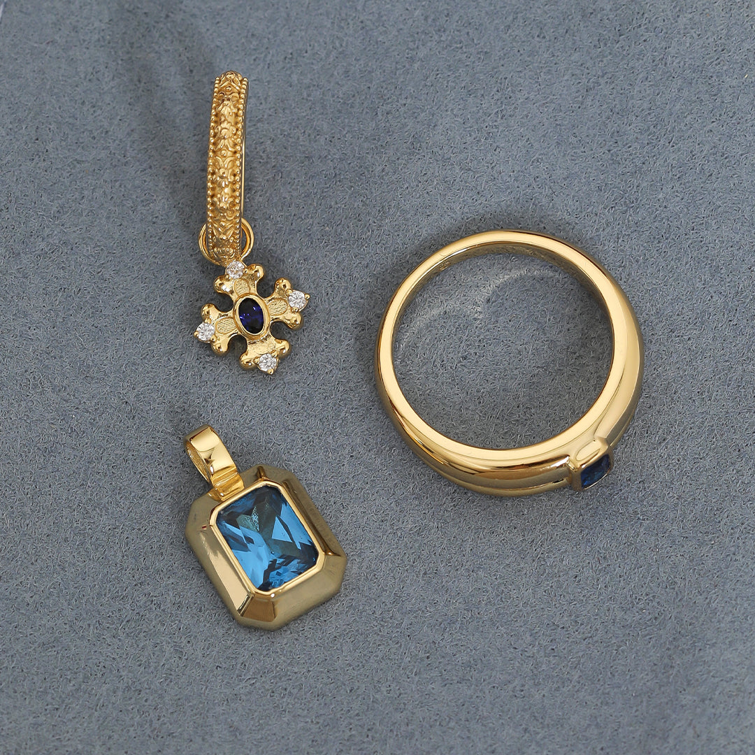 gold charm that features the intricate fleur-de-lis details found in the iconic Palace of Versailles accompanied by the deep blue stone on gold hoops next to charm and ring on navy background